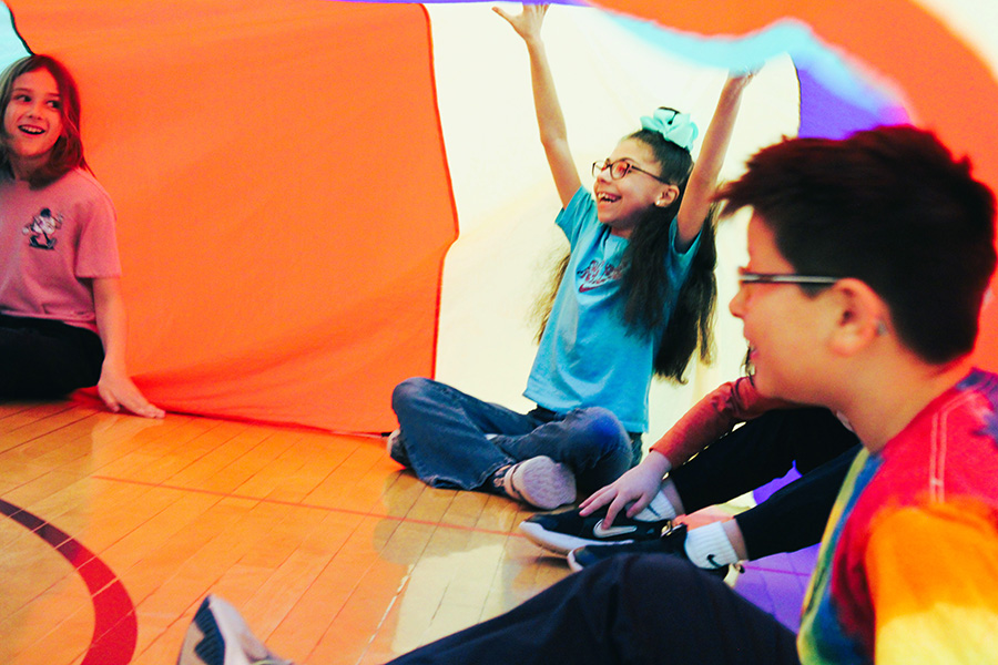 Third grade students play inside rainbow parachute during gym class.
