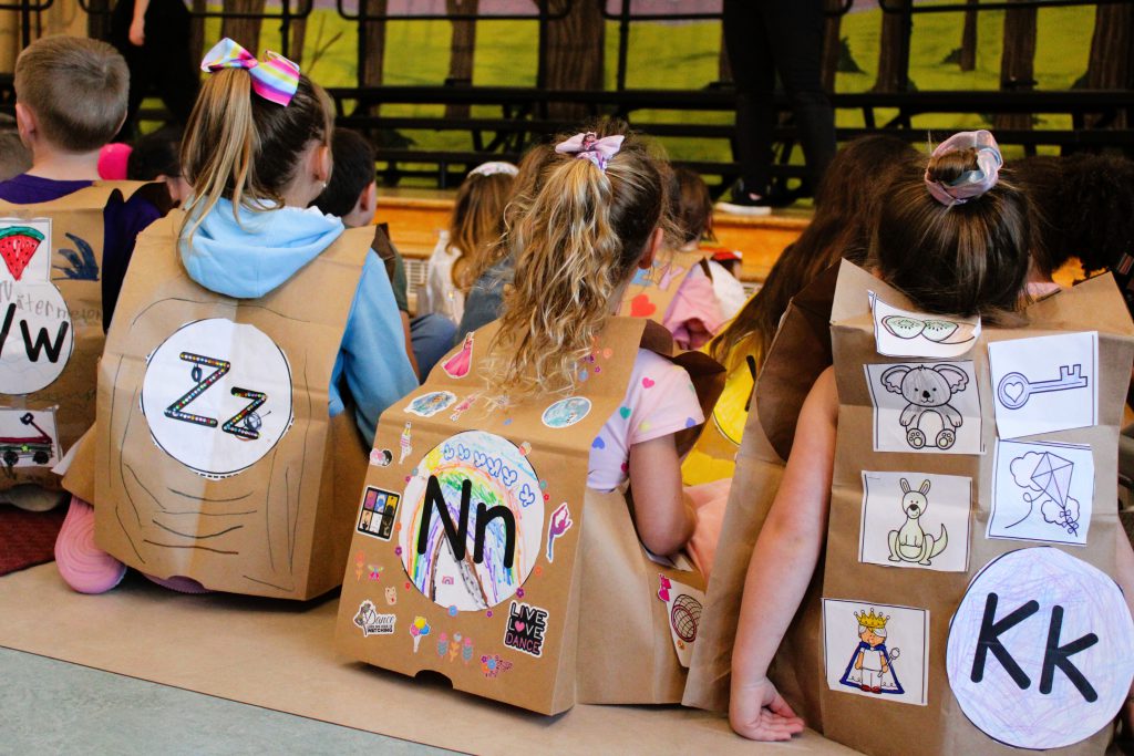 Tecumseh Kindergarten students sit on floor wearing ABC fashion show paper vests with letters.