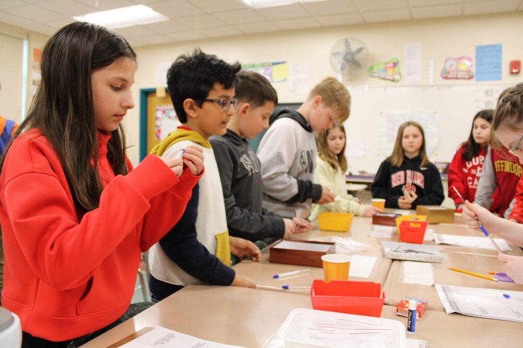 Fourth grade students stand in a line along desks helping build pens.
