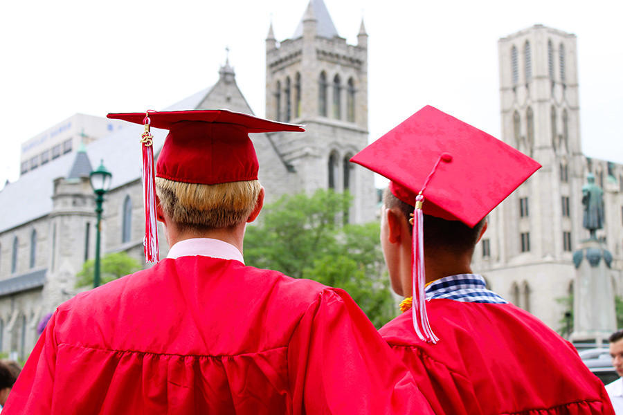 View from behind two graduates wearing red caps and gowns with the city in the background.