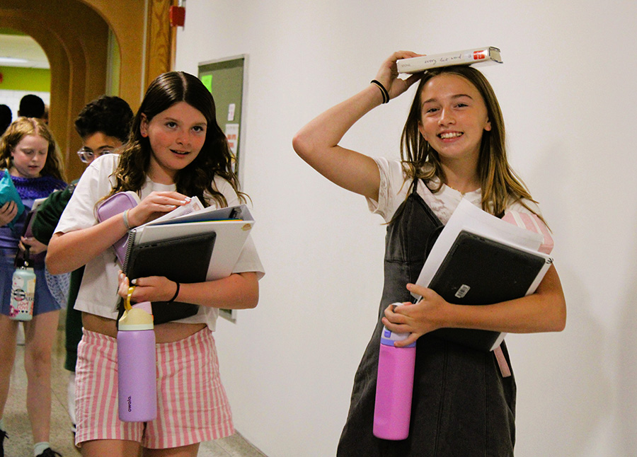 Student balances book on head, smiling at camera while walking to class.