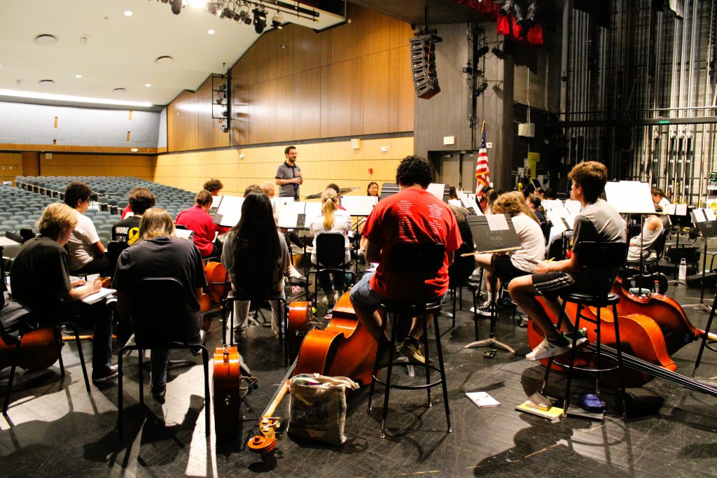 Orchestra students listen to conductor on stage.