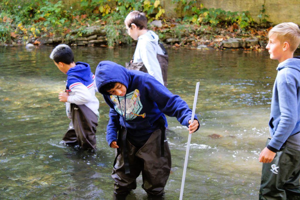 Student wade through stream with measuring tools.