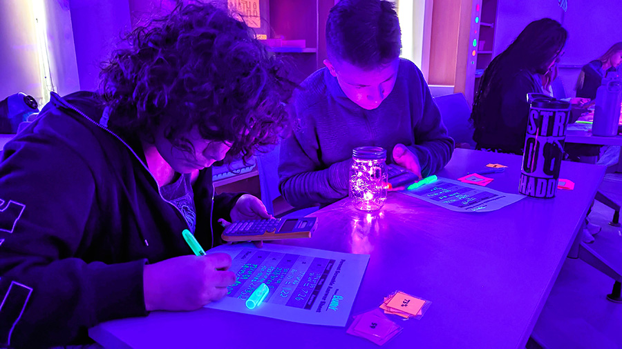 Two students sit at desk doing math work in black light lighting.