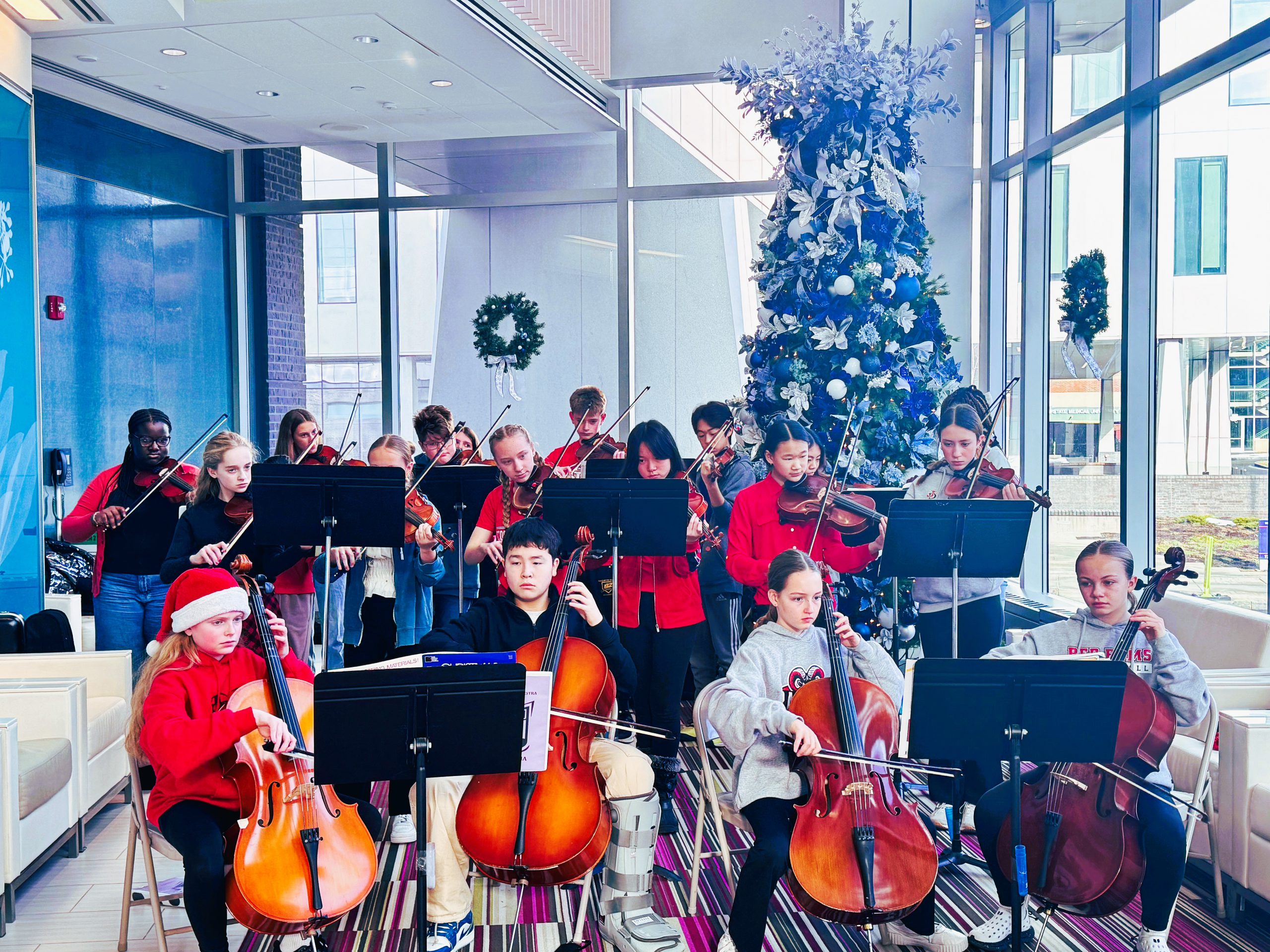 Middle school orchestra students perform in holiday decorated hospital area.