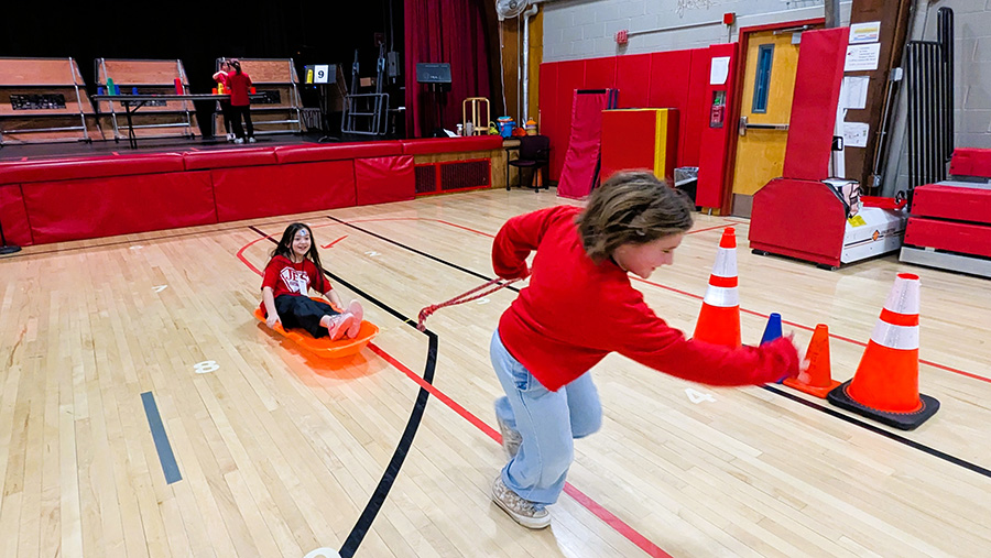 Student pulls another in a sled in school gym during winter carnival.