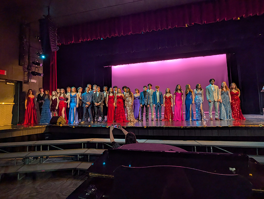 Students stand in line on stage wearing prom dresses and tuxedos. 