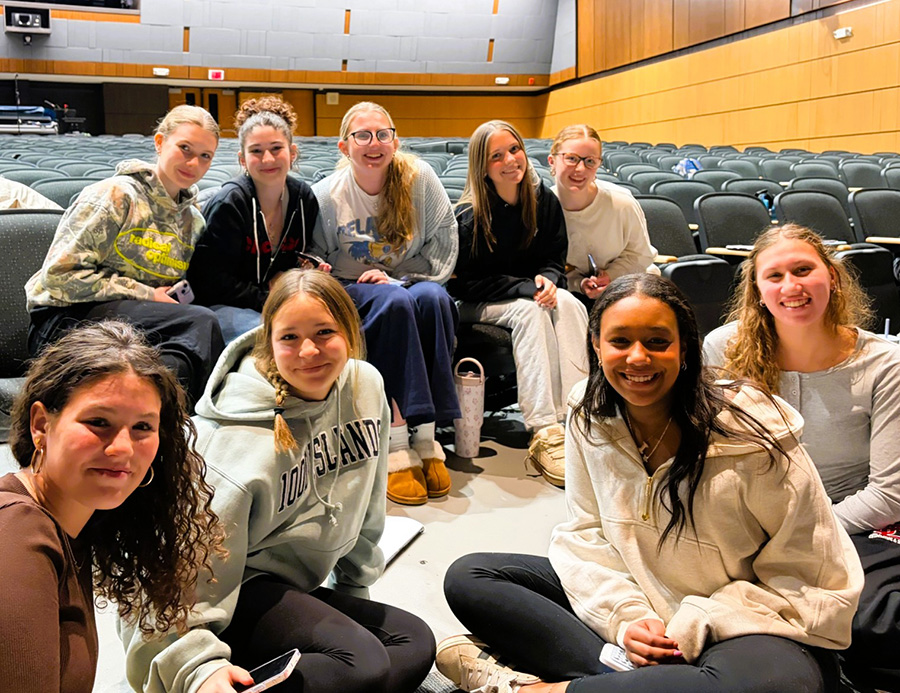 Students pose for group photo sitting in chairs and floor of auditorium.