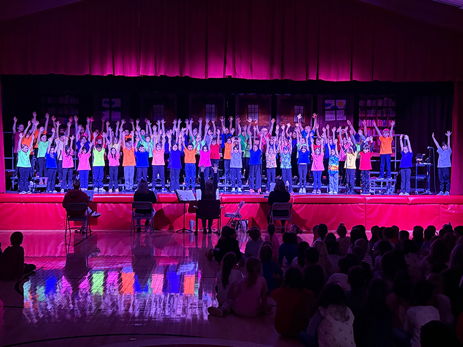 Students in colorful T-shirts stand with arms raised on stage in front of crowd.