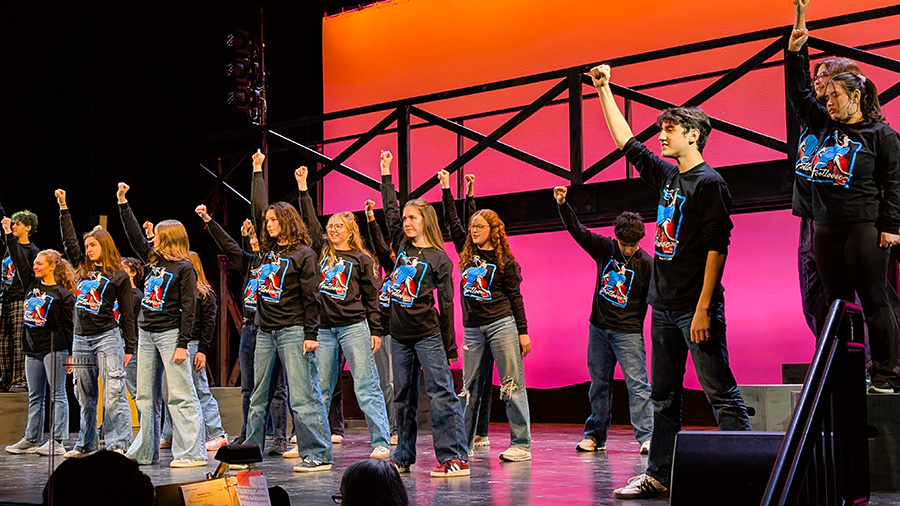 Students perform on stage wearing black Footloose promotion shirts with one fist in the air.