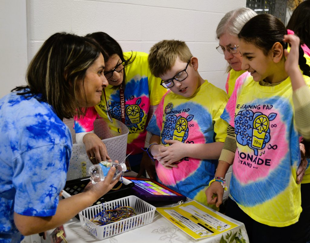 Two students and two staff members work lemonade stand, smile at customer holding bottle.