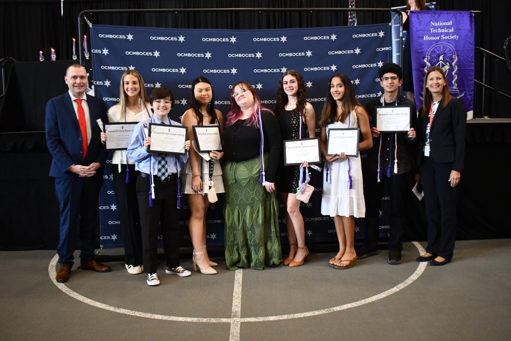 Eight students pose for group photo holding certificates with the principal on the left and superintendent on the right. An OCM BOCES background is behind them.