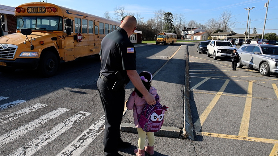School Patrol Officer stands with students with backs to the camera as they watch buses drive by.