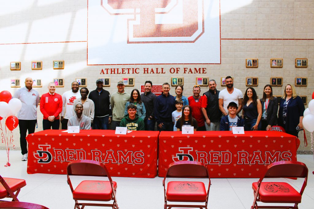 Four student-athletes sit at table while coaches, family, and admin stand behind them for group photo.