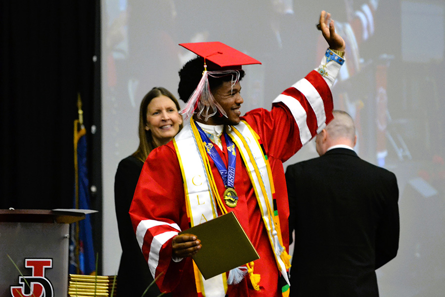 Graduate waves hand at audience with diploma in hand.
