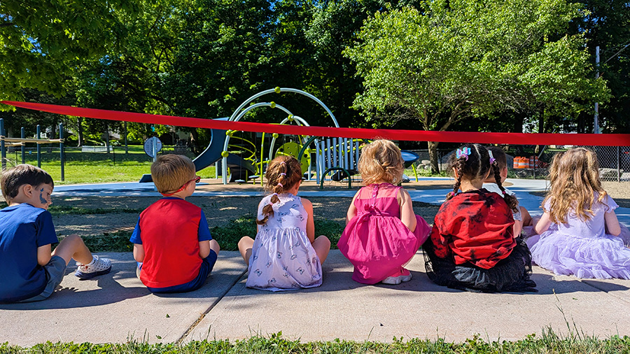 UPK students sit on ground next to ribbon with new playground in the background.