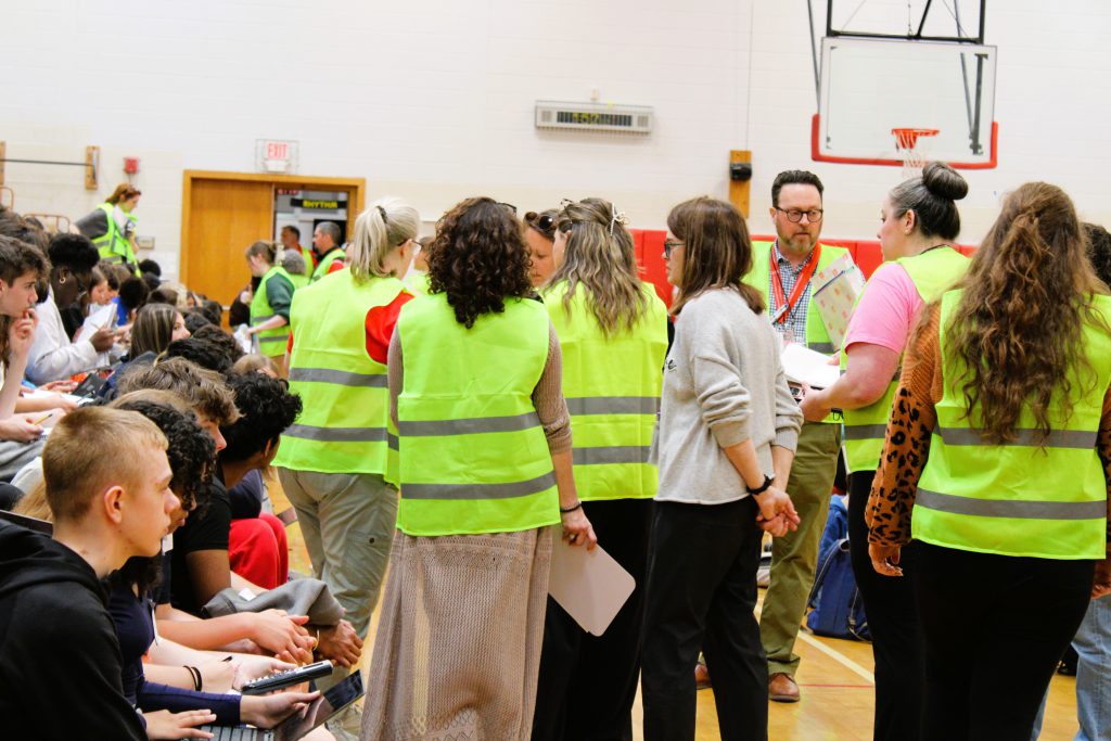 Staff stand in gym wearing yellow vests talking in gym full of students.