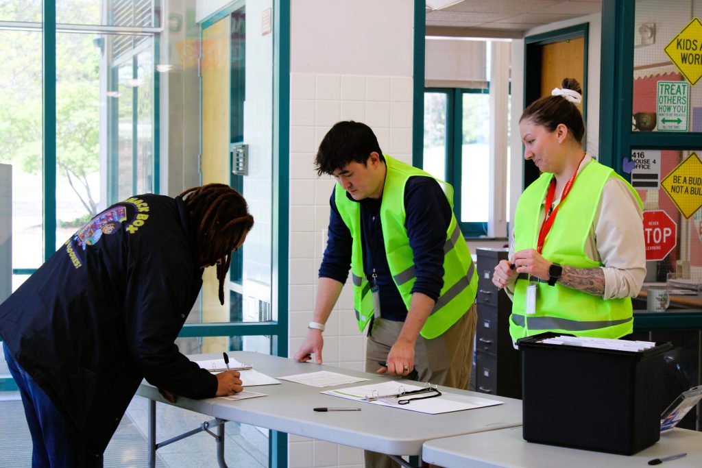 Two staff members wearing yellow vests check parent in during drill.