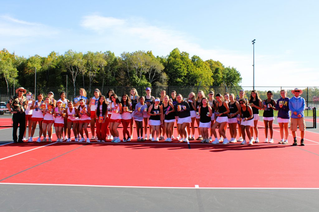 J-D and CBA Girls Tennis teams pose for group photo on tennis court while holding food drive donations.